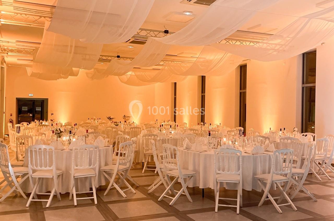 Salle de réception décorée avec des tables rondes dressées, des chaises blanches et un éclairage chaleureux.