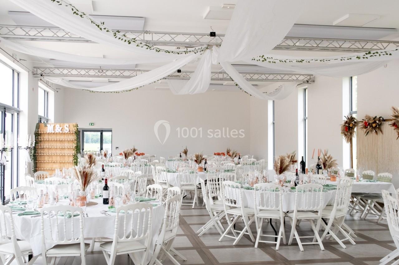 Salle de réception lumineuse décorée pour un événement, avec tables rondes dressées et ornements floraux.