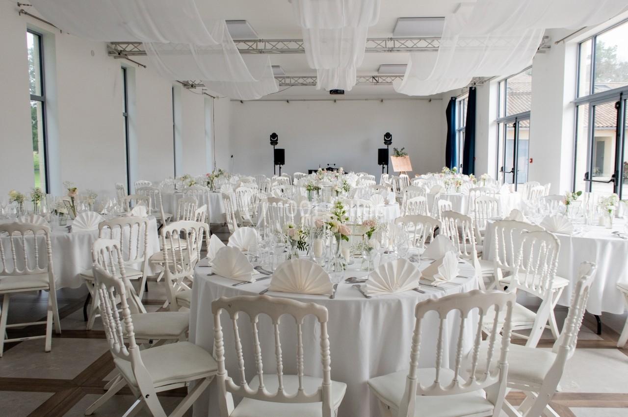 Salle de réception lumineuse avec des tables rondes décorées de nappes blanches, fleurs et pliages de serviettes.