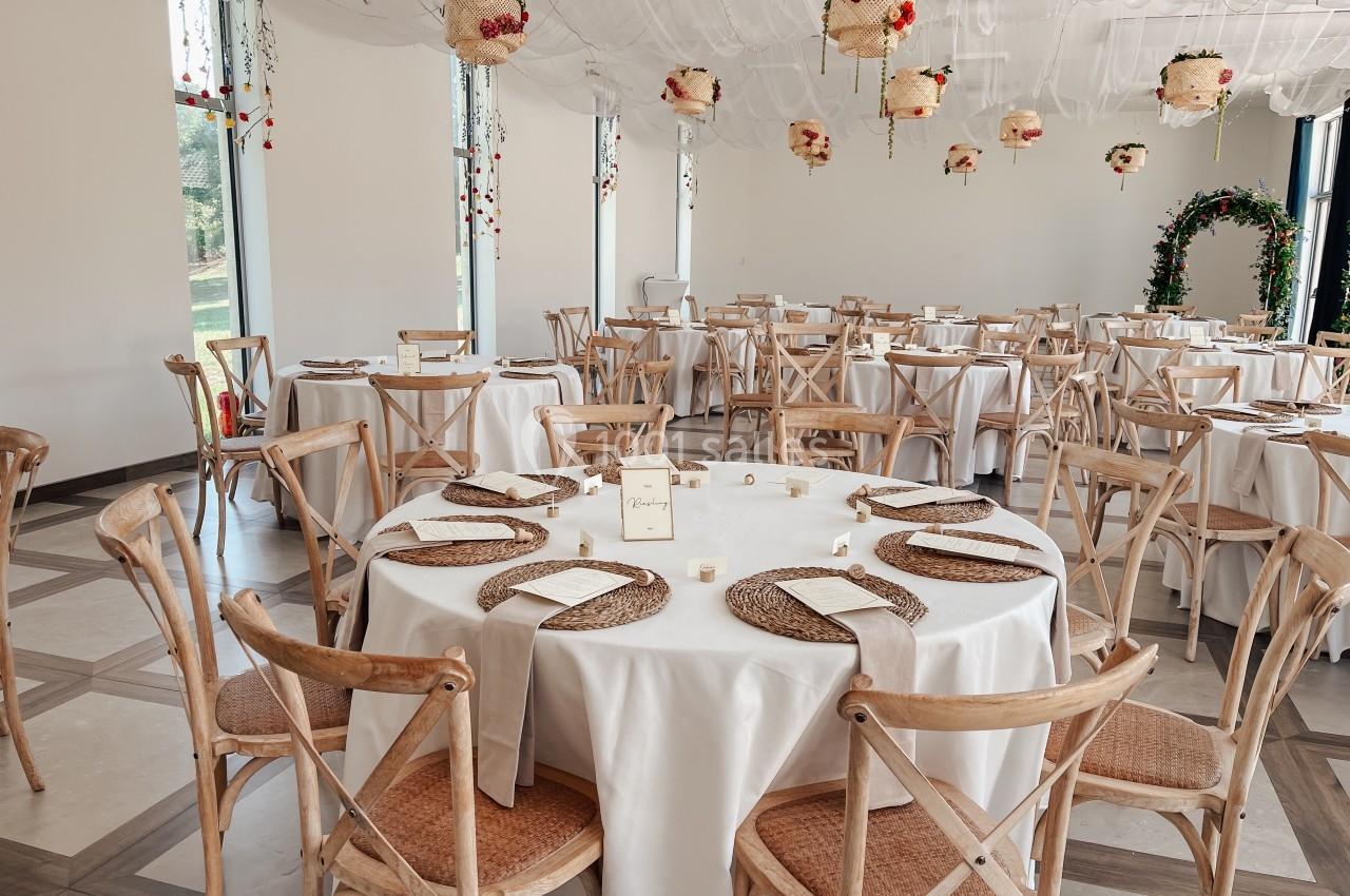 Salle de réception décorée avec des tables rondes dressées, des chaises en bois et des suspensions florales.