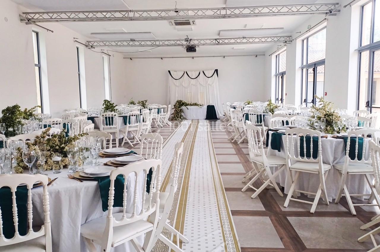 Salle de réception lumineuse avec tables rondes décorées, chaises blanches et allée centrale bordée de motifs.