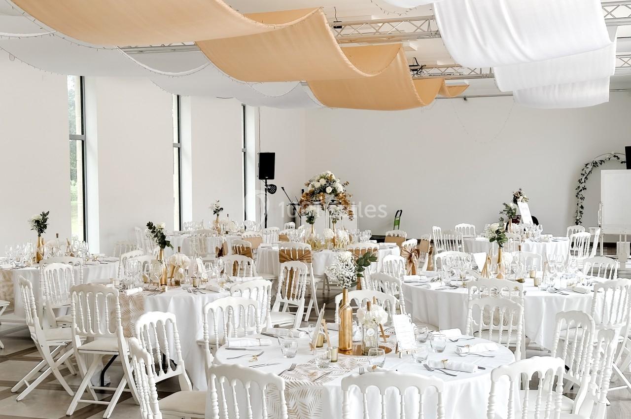 Salle de réception décorée avec des tables rondes dressées, des chaises blanches et des voilages suspendus au plafond.