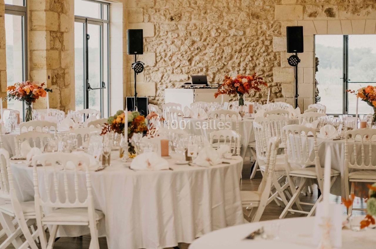 Salle de réception lumineuse avec tables rondes dressées, chaises blanches et lustres suspendus au plafond en bois.