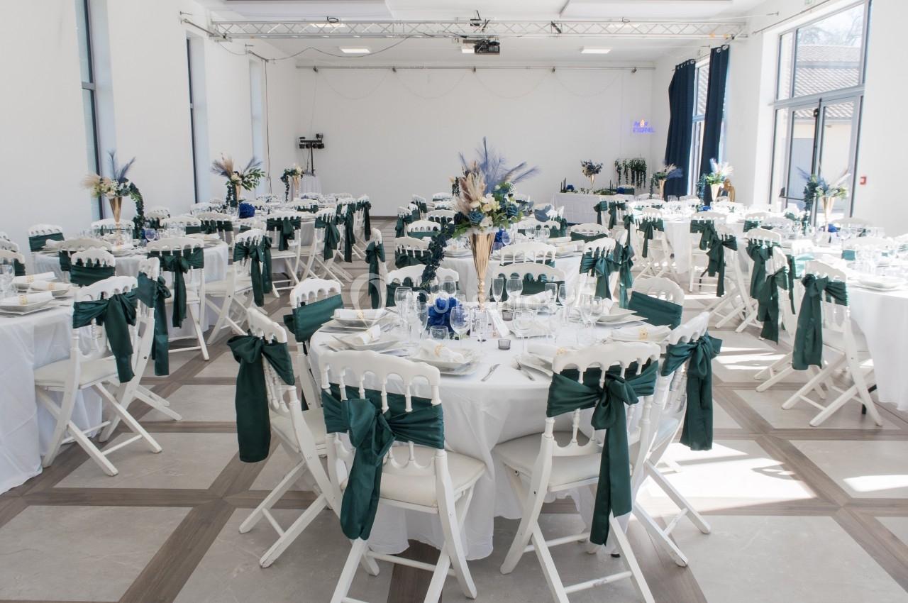 Salle de réception lumineuse avec tables rondes dressées, décorées de nappes blanches et de chaises ornées de nœuds verts.