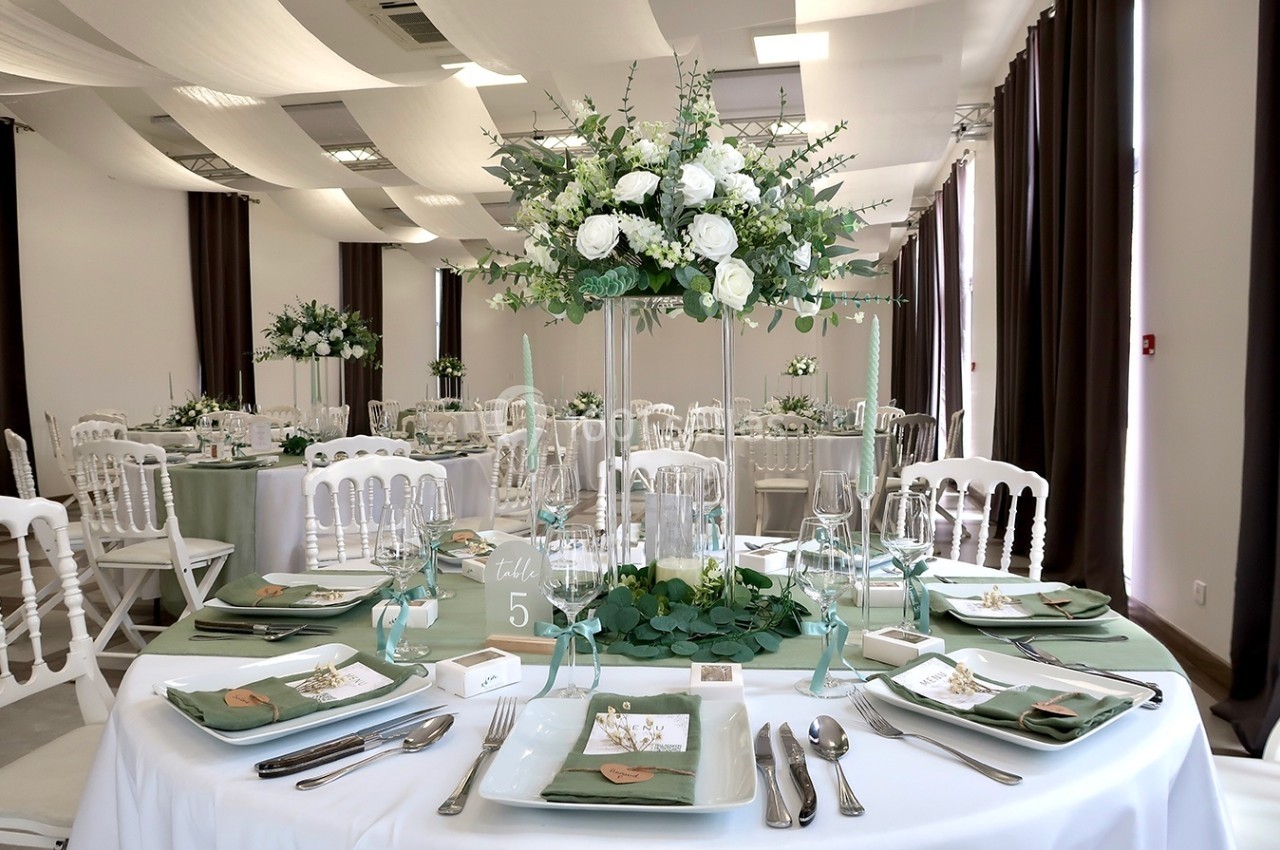Salle de réception décorée pour un mariage, avec tables dressées, nappes blanches, centres de table fleuris et chaises…