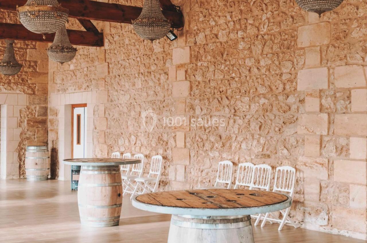 Salle en pierre avec des poutres apparentes, des lustres suspendus, des tonneaux en bois et des chaises blanches alignées.