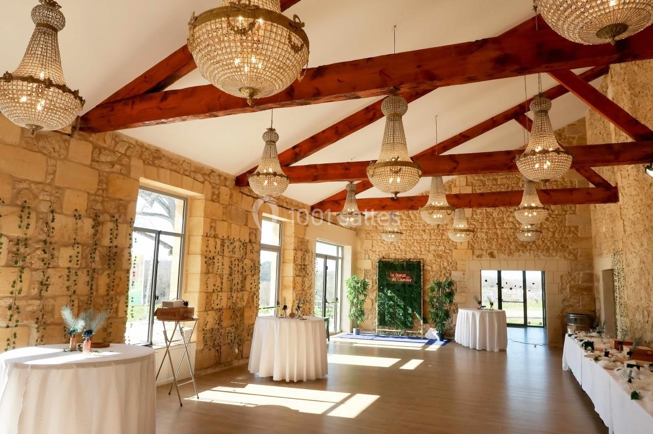 Salle lumineuse avec murs en pierre, poutres apparentes, lustres en cristal et tables décorées pour un événement.