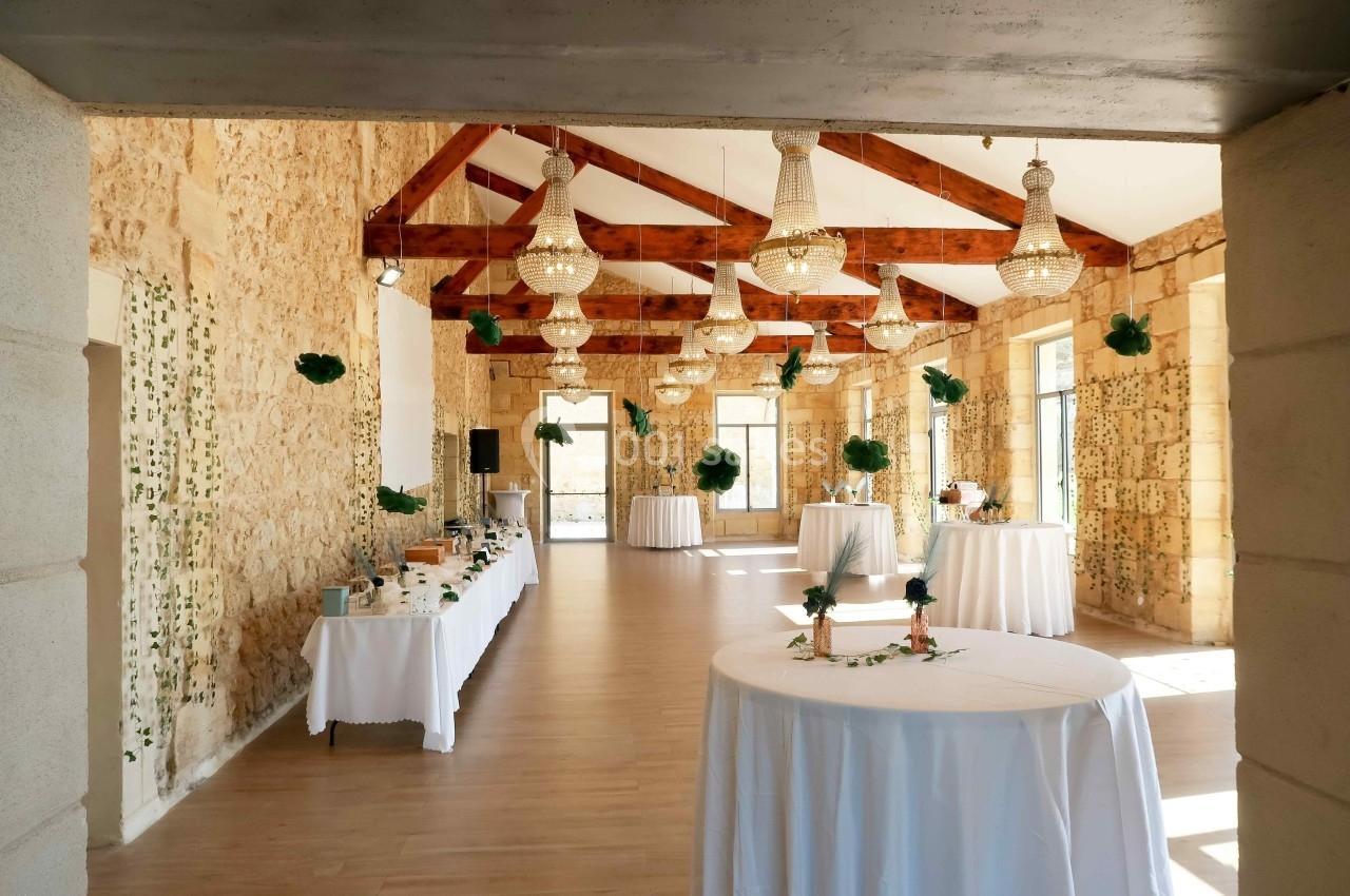 Salle de réception lumineuse avec murs en pierre, poutres apparentes, tables décorées et lustres suspendus.