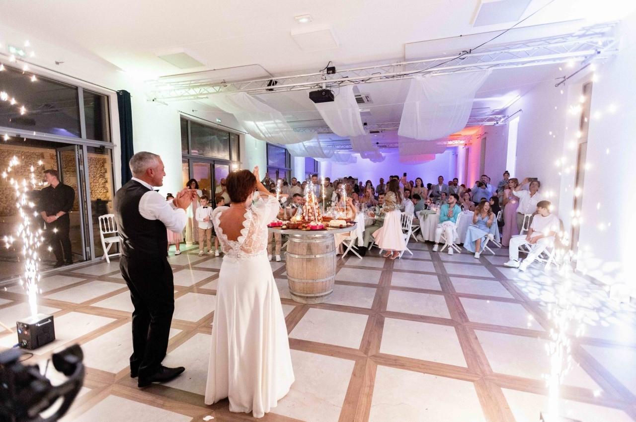 Un couple célèbre son mariage devant des invités dans une salle décorée, avec des étincelles et une ambiance festive.