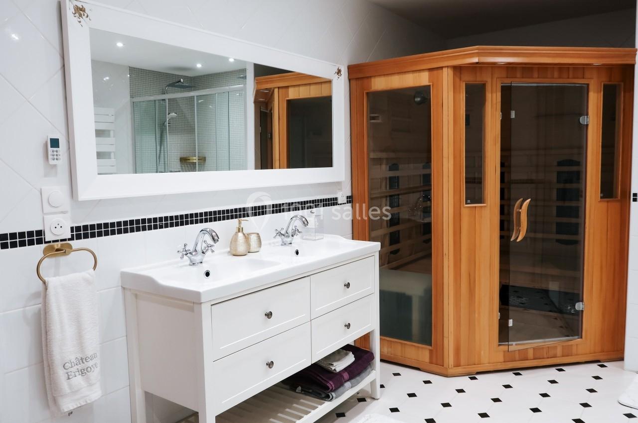 Salle de bain avec double vasque blanche, miroir mural, sauna en bois et carrelage noir et blanc.