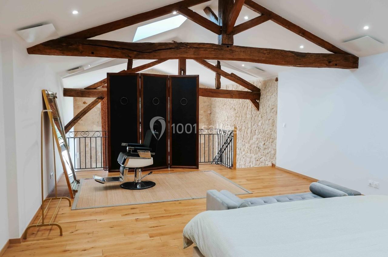 Aménagement d'une pièce lumineuse avec poutres apparentes, fauteuil de barbier, miroir sur pied et lit au premier plan.