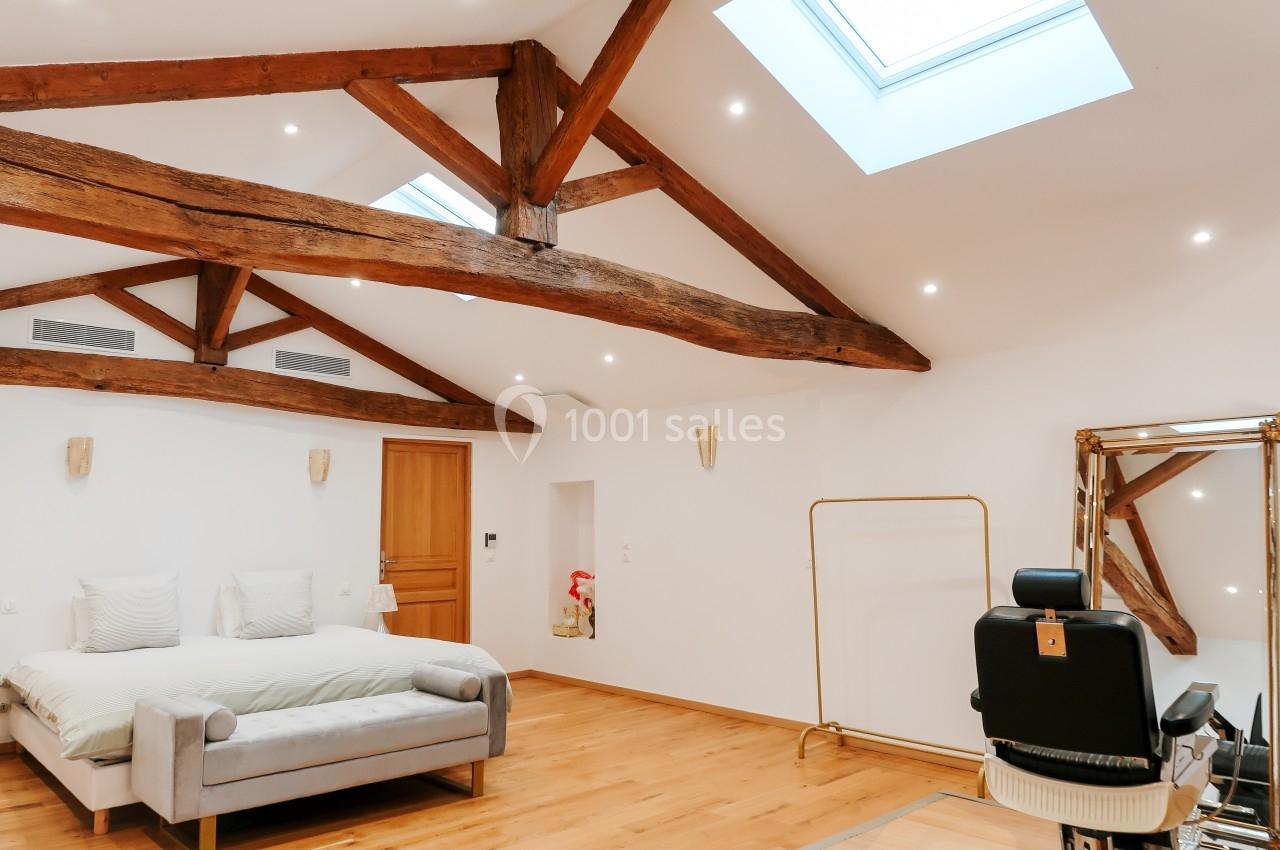 Chambre lumineuse avec poutres apparentes, lit double, fauteuil de coiffure, miroir et parquet en bois clair.