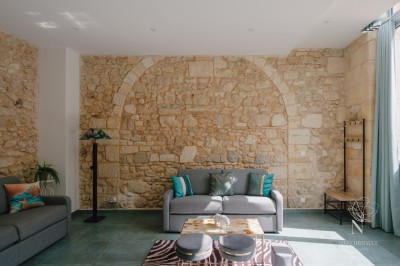 Salon lumineux avec murs en pierre apparente, canapé gris, table basse et grandes fenêtres laissant entrer la lumière.