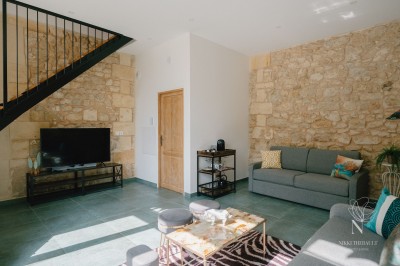 Salon lumineux avec murs en pierre apparente, canapé gris, table basse et grandes fenêtres laissant entrer la lumière.