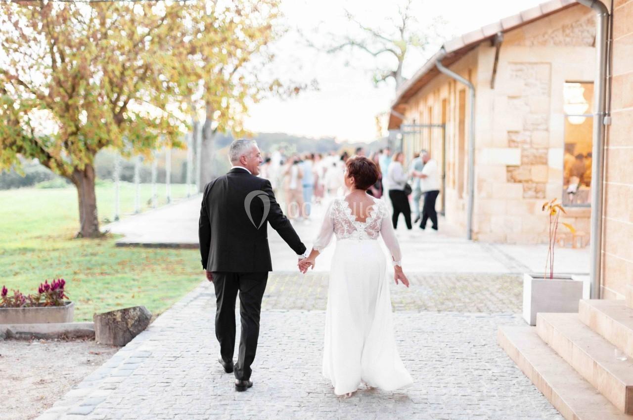Un couple en tenue de mariage marche main dans la main sur une allée pavée, près d'un bâtiment en pierre.