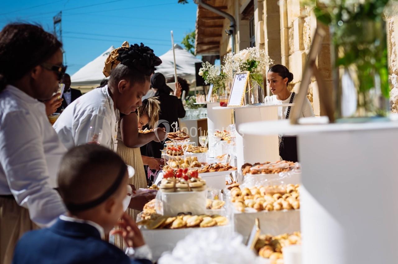Des personnes se servent à un buffet en extérieur, avec des tables décorées de fleurs et des pâtisseries variées.