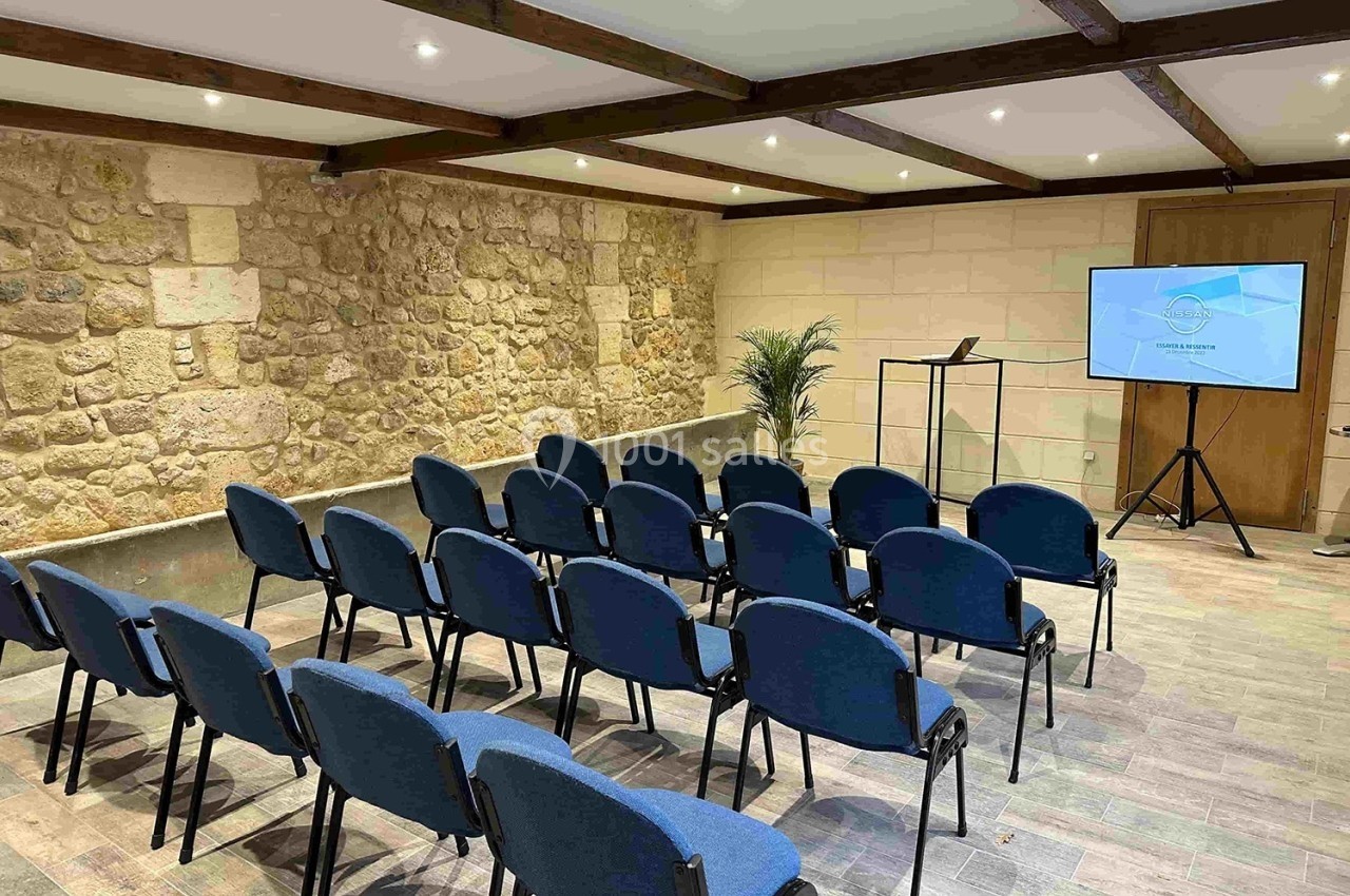 Salle de conférence avec des chaises bleues alignées, un écran de présentation et un pupitre devant un mur en pierre.
