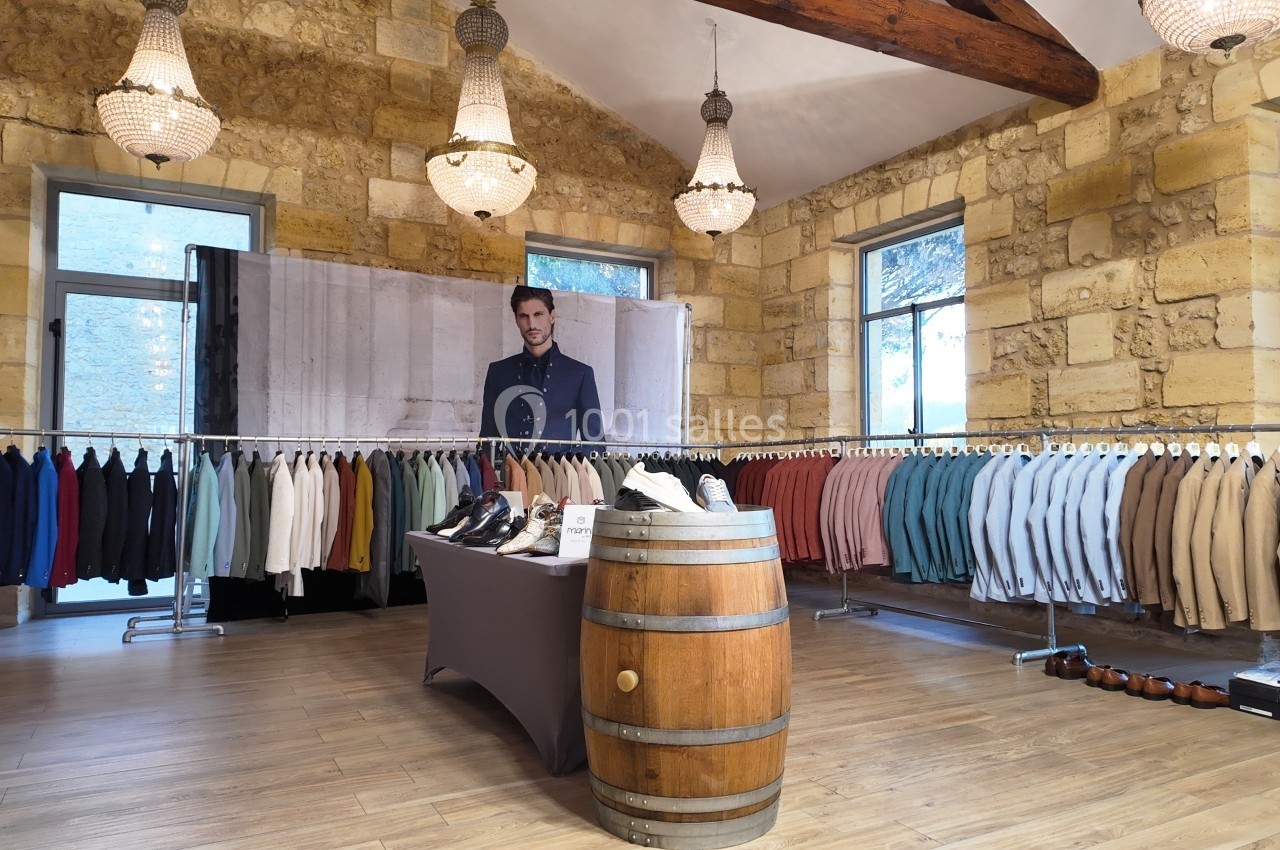 Salle avec des vêtements suspendus sur des portants, chaussures exposées sur une table et un tonneau, murs en pierre…