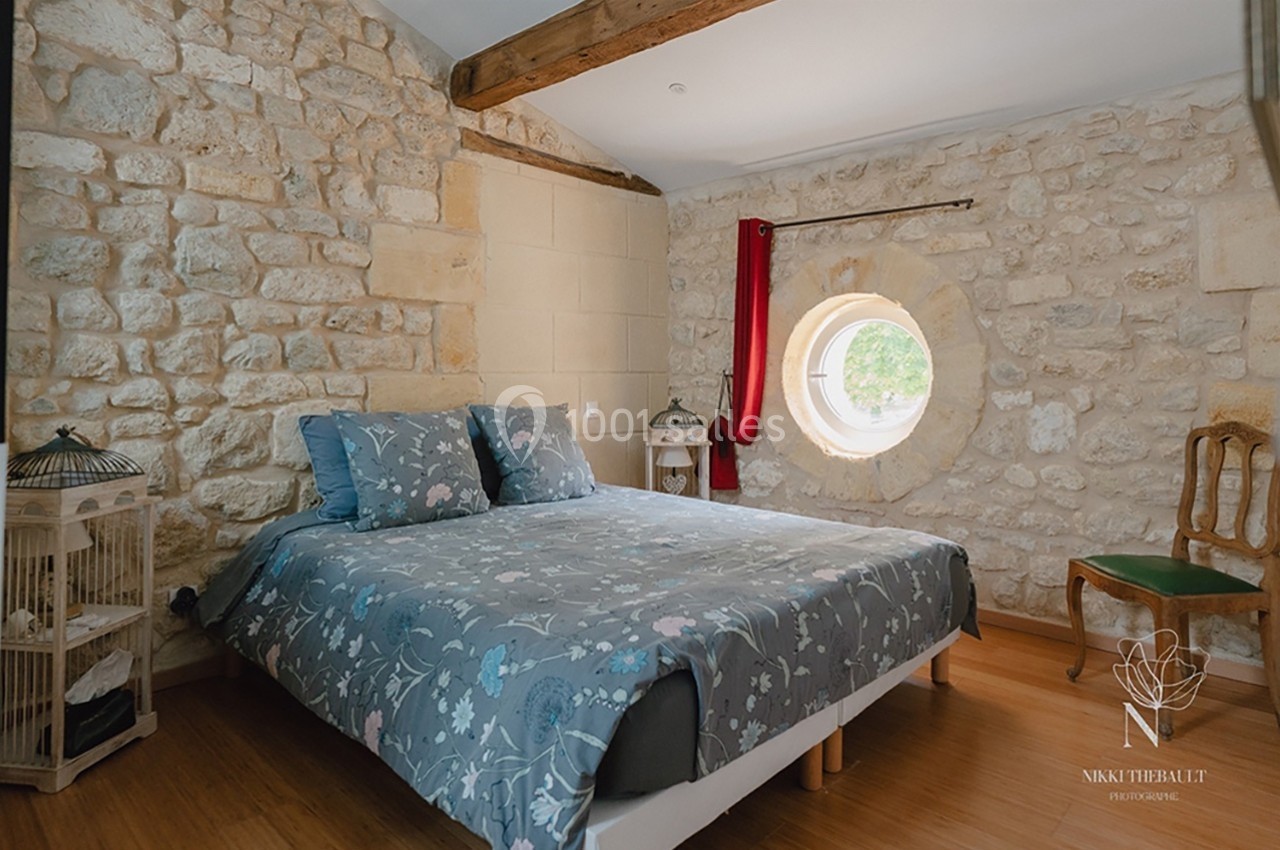 Chambre avec murs en pierre apparente, lit double avec couvre-lit fleuri, fenêtre ronde et poutres en bois au plafond.