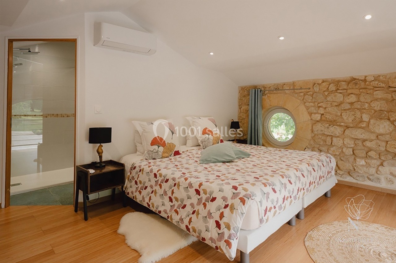 Chambre lumineuse avec lit double, murs en pierre, fenêtre ronde, parquet et salle de bain attenante visible.