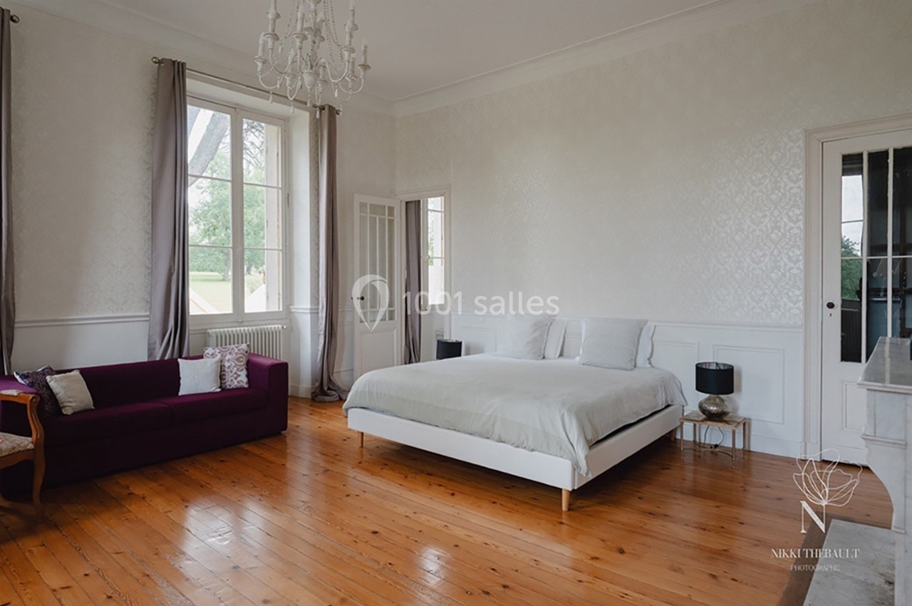 Chambre lumineuse avec lit double, canapé violet, parquet en bois, grandes fenêtres et murs clairs.