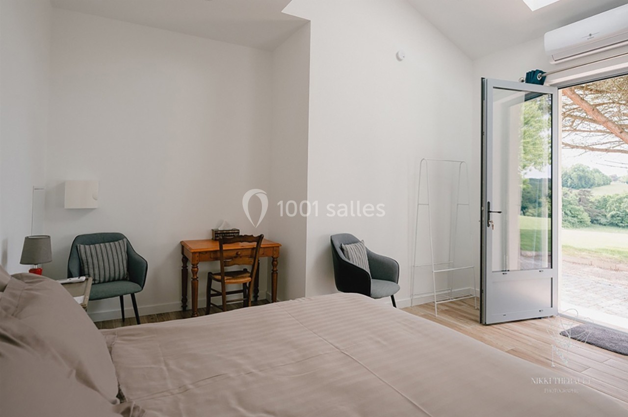 Chambre lumineuse avec lit double, bureau en bois, deux fauteuils et porte-fenêtre ouverte sur un jardin verdoyant.