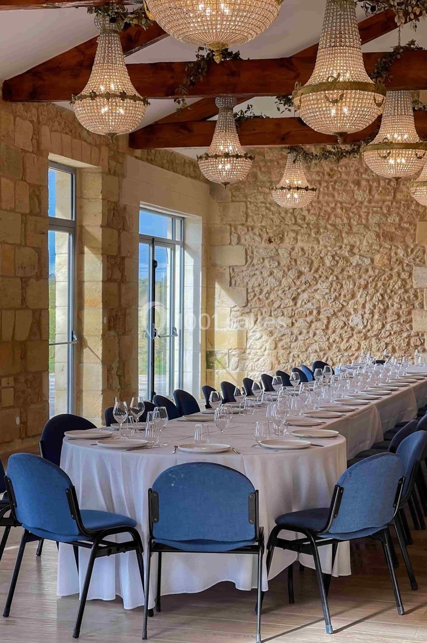 Salle lumineuse avec murs en pierre, grandes fenêtres, lustres en cristal et tables rondes dressées avec nappes blanches.