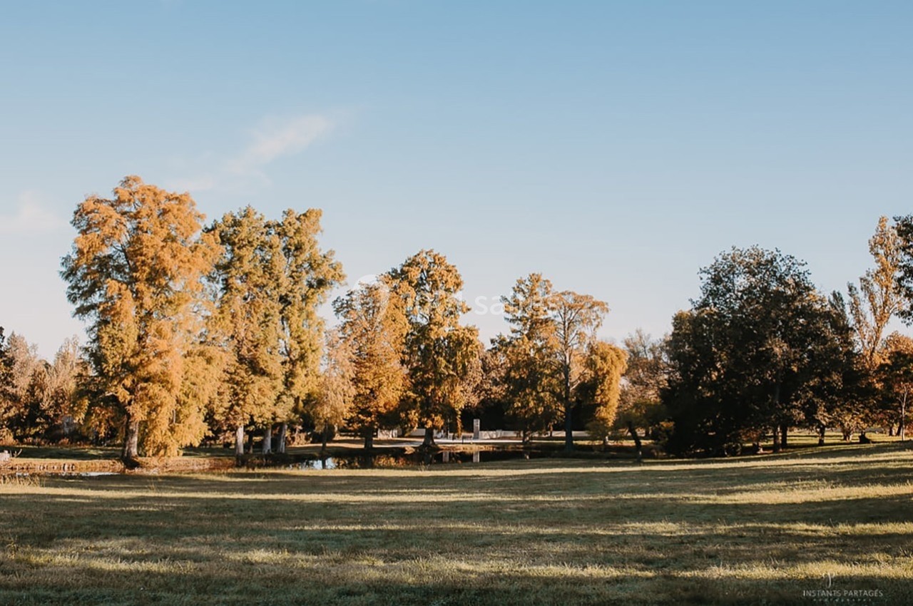 Pelouse verdoyante bordée d'arbres aux teintes automnales, avec un petit pont surplombant un étang en arrière-plan.