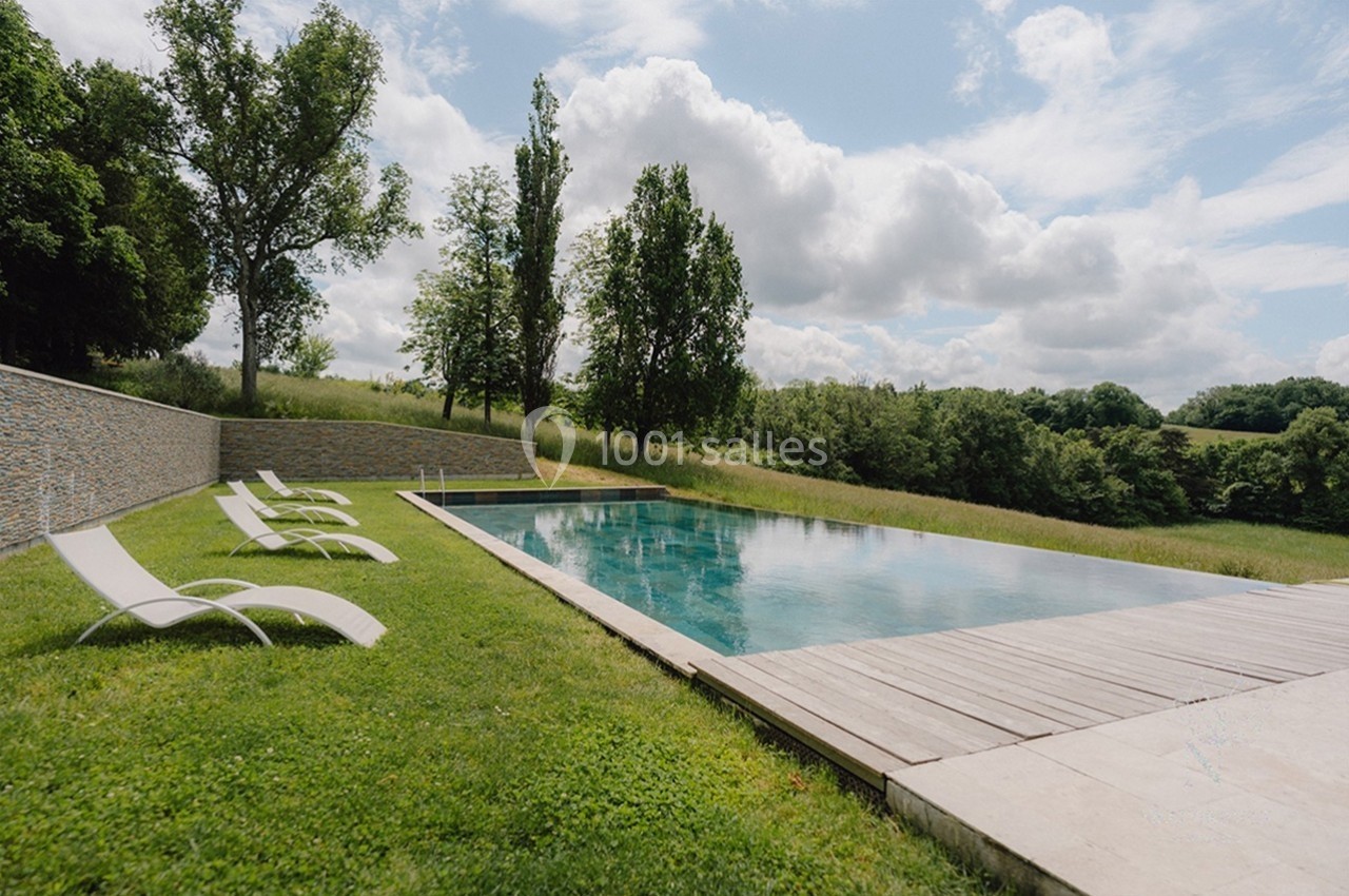 Piscine extérieure à débordement entourée de chaises longues, située dans un cadre verdoyant sous un ciel nuageux.