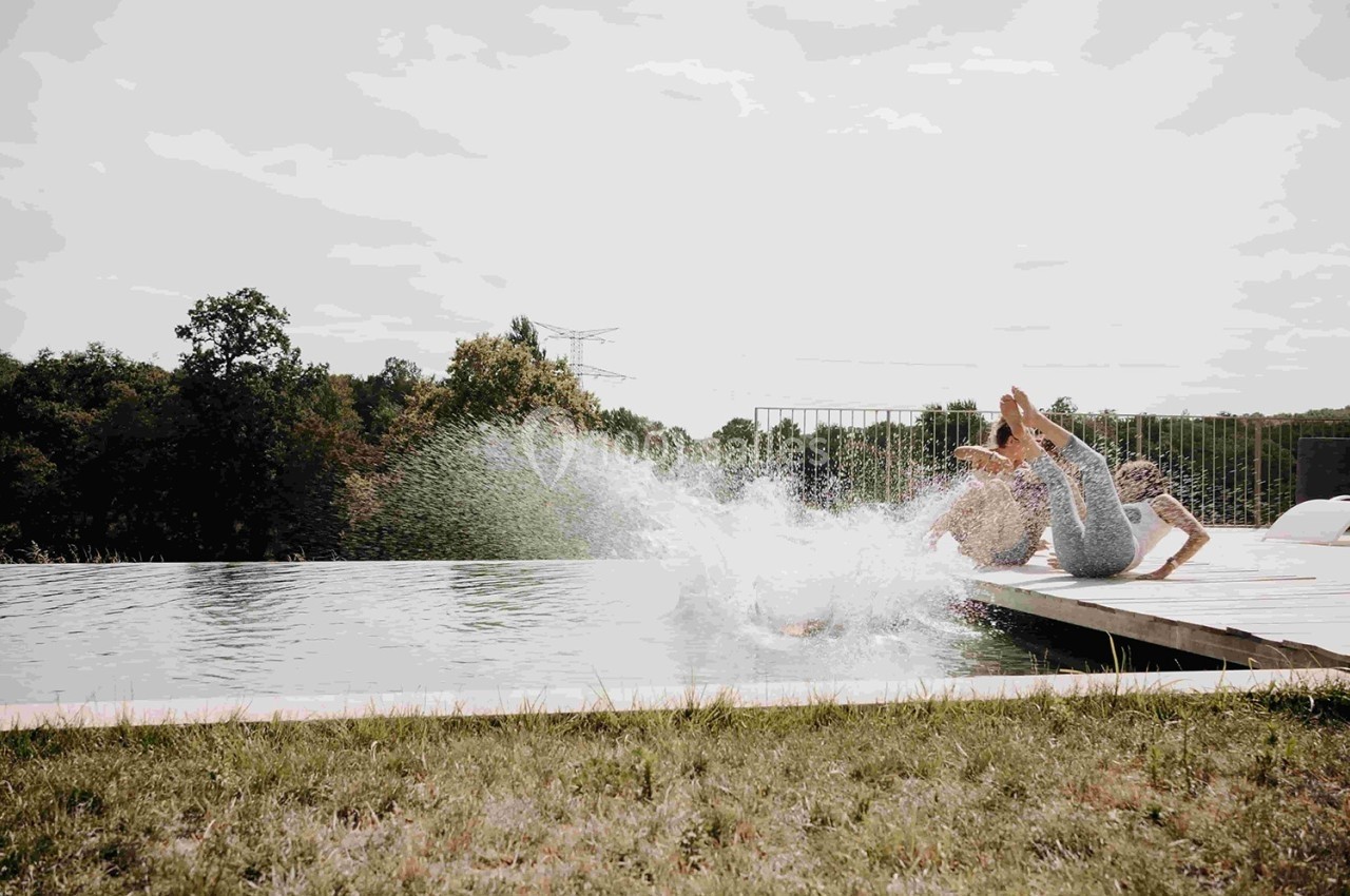 Deux personnes plongent dans une piscine depuis un ponton en bois, entourée de nature verdoyante.