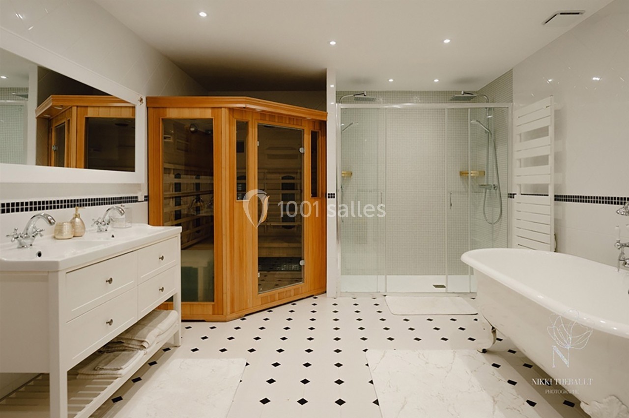 Salle de bain moderne avec sauna en bois, douche vitrée, baignoire sur pieds et double vasque sur meuble blanc.