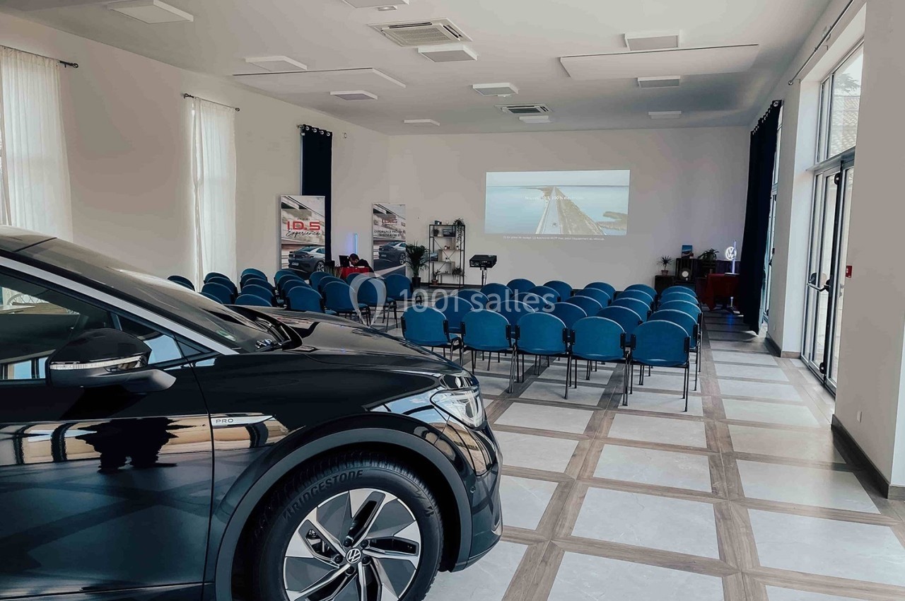 Salle de conférence lumineuse avec des rangées de chaises bleues, un écran de projection et une voiture noire au premier…
