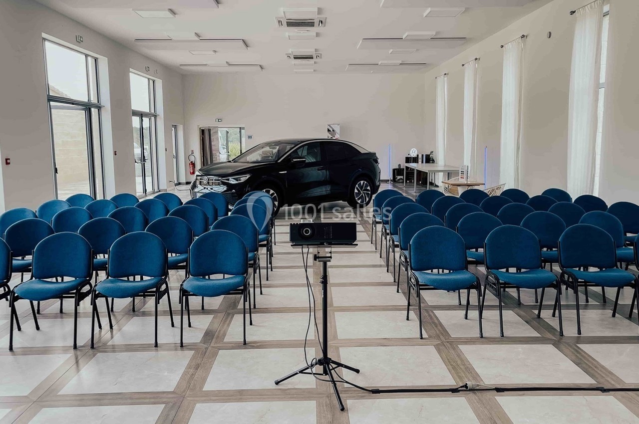 Salle de conférence lumineuse avec des rangées de chaises bleues et une voiture exposée au fond.