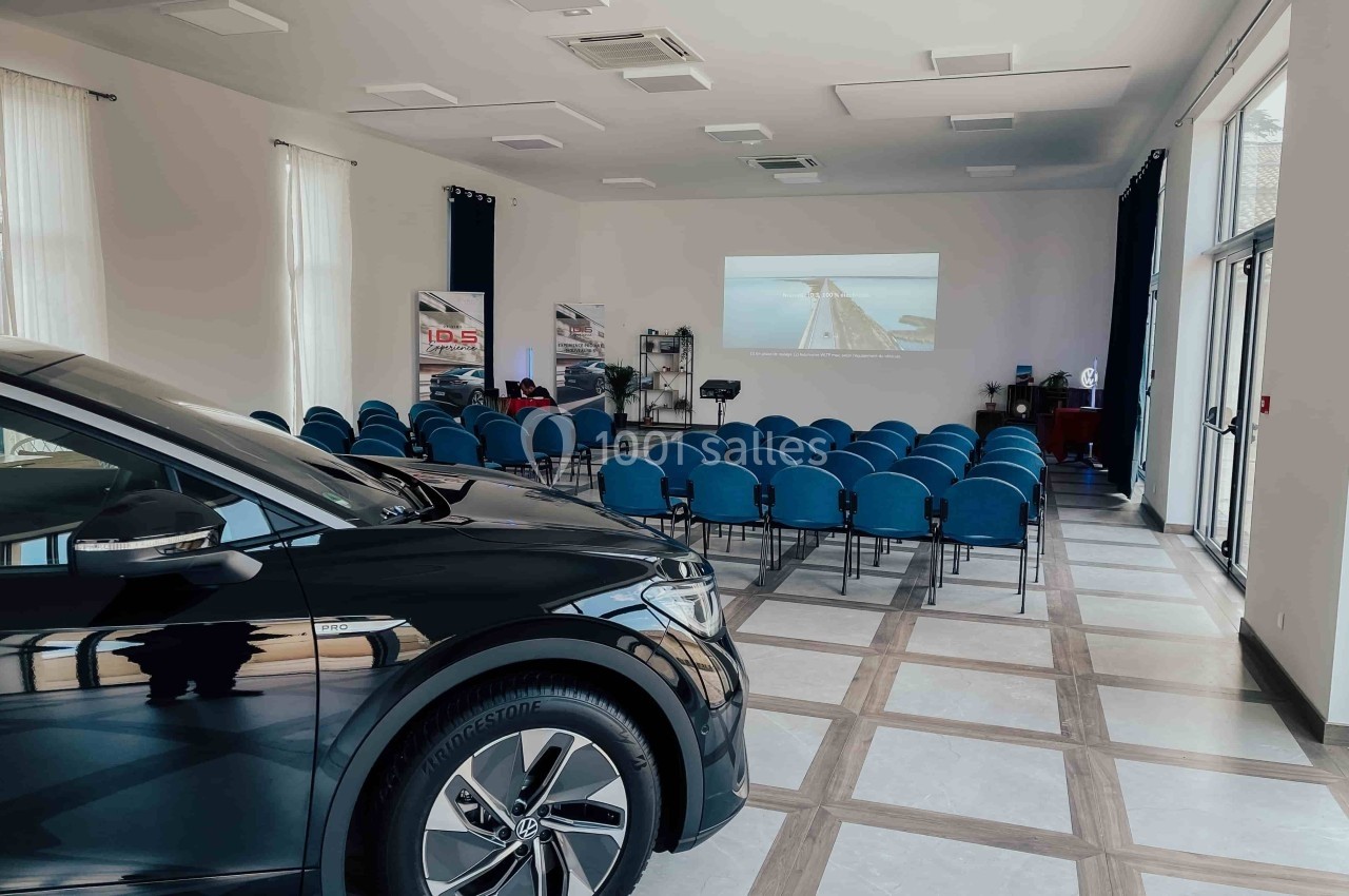 Salle de conférence lumineuse avec des rangées de chaises bleues, un écran de projection et une voiture au premier plan.