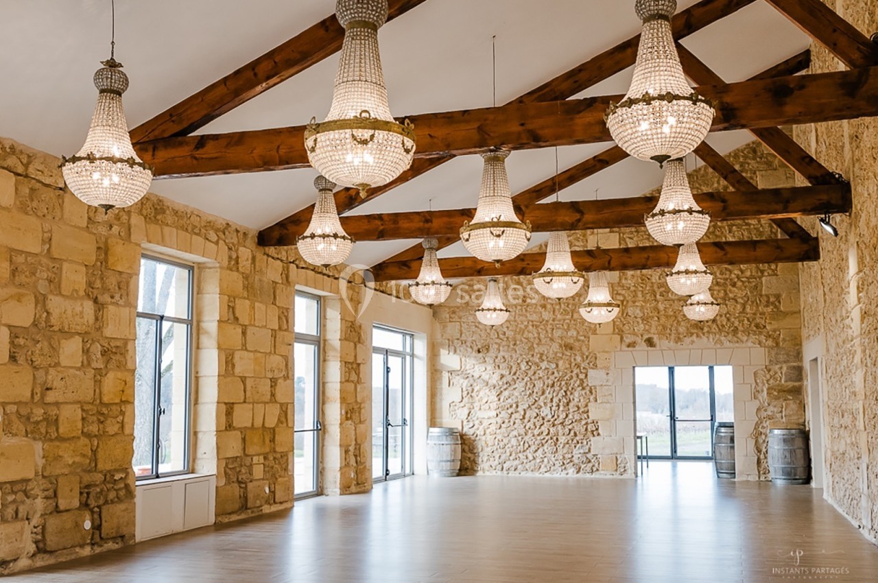 Salle spacieuse avec murs en pierre, poutres apparentes et lustres suspendus, éclairée par de grandes fenêtres.