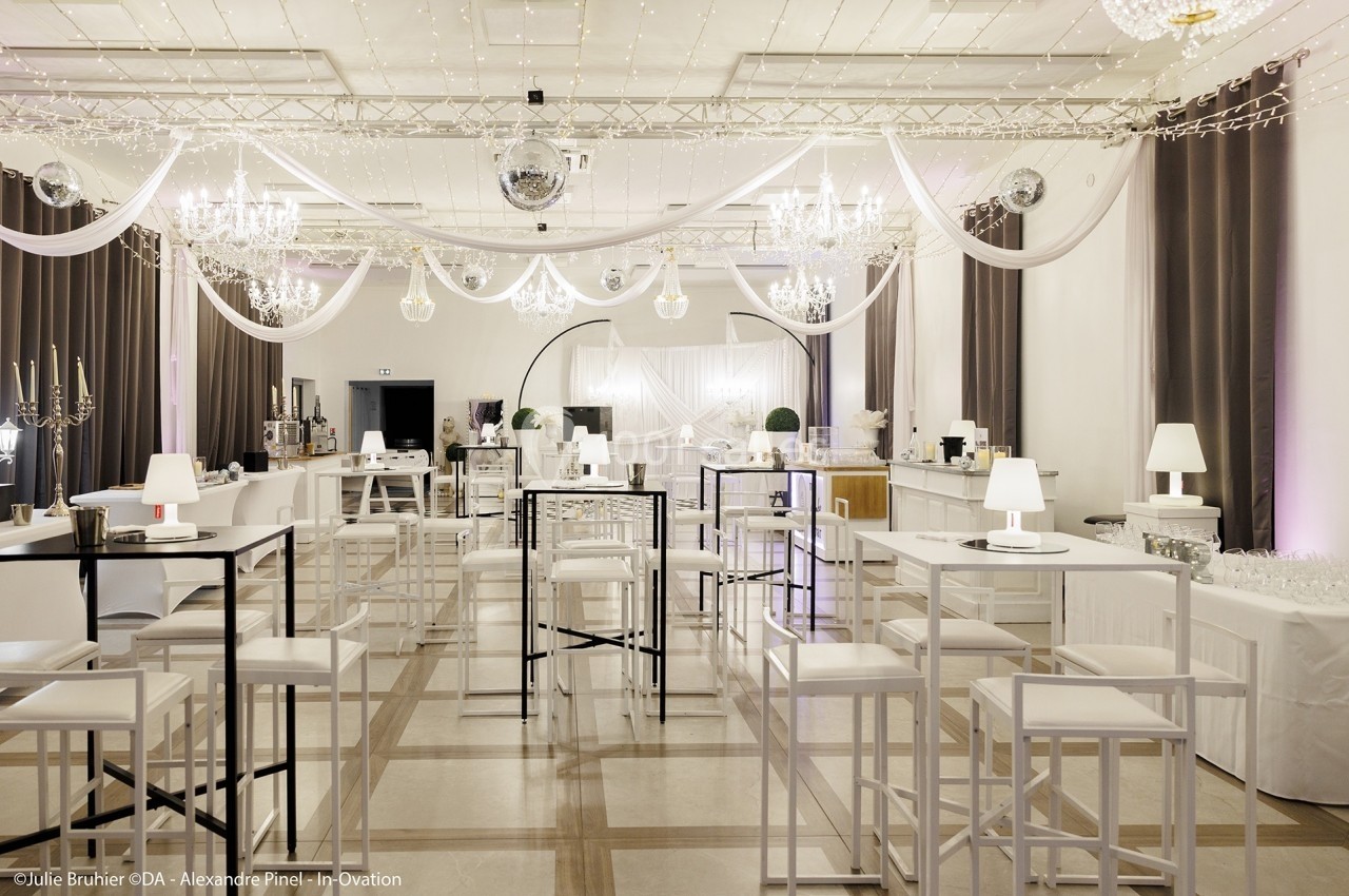 Salle de réception décorée avec des tables hautes, des guirlandes lumineuses et des lustres suspendus.