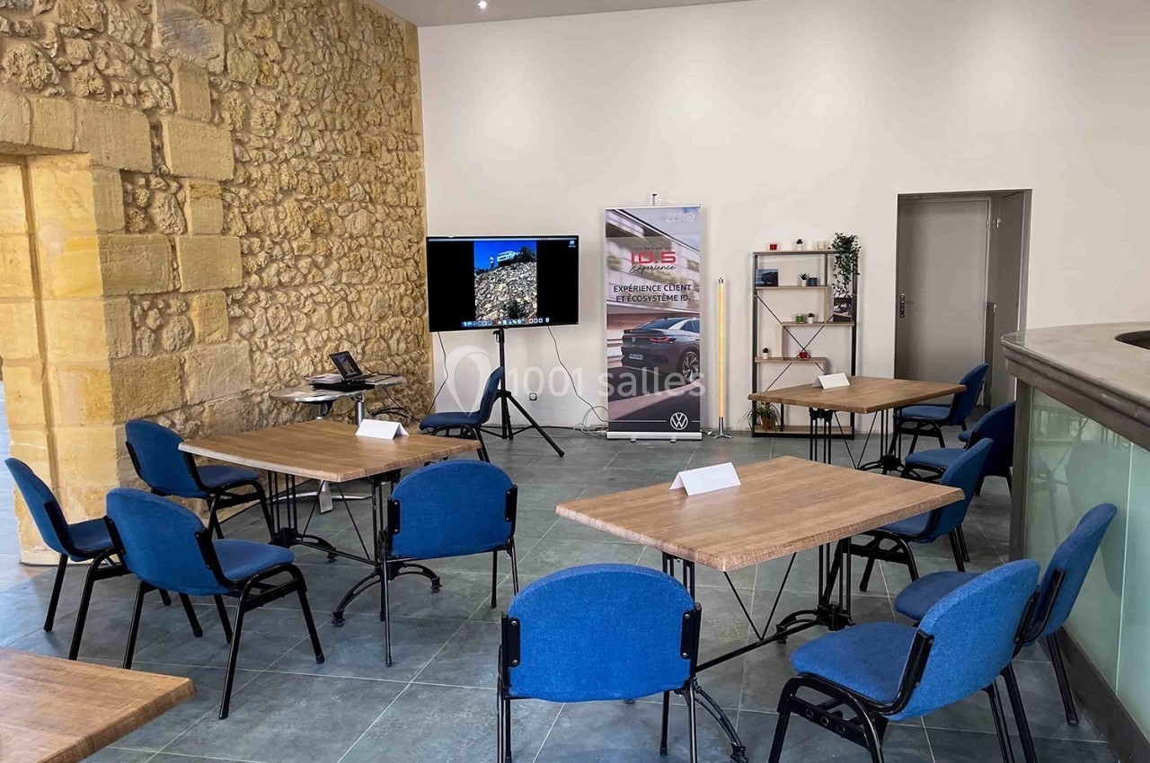 Salle de réunion avec murs en pierre, tables en bois, chaises bleues et écran de présentation.