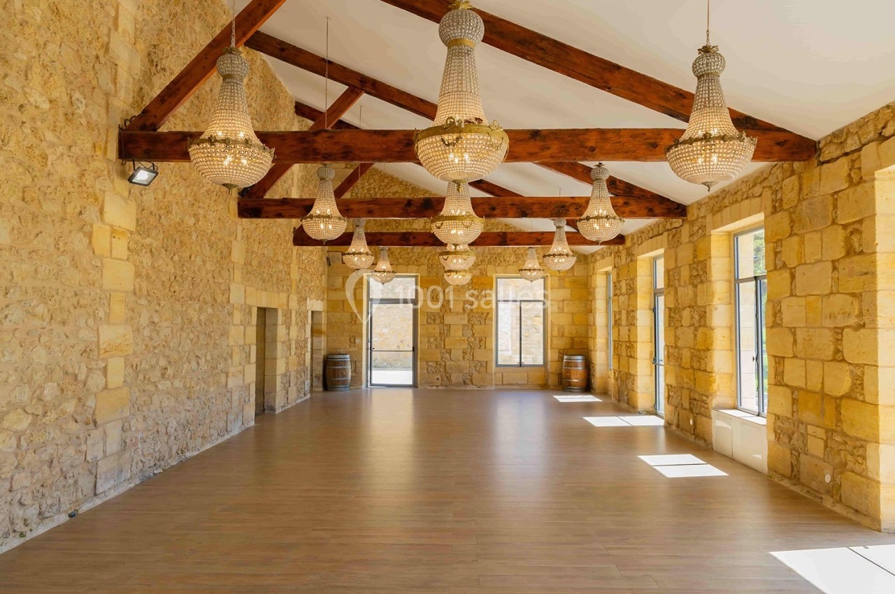 Salle lumineuse avec murs en pierre, poutres apparentes et lustres en cristal suspendus, parquet au sol.