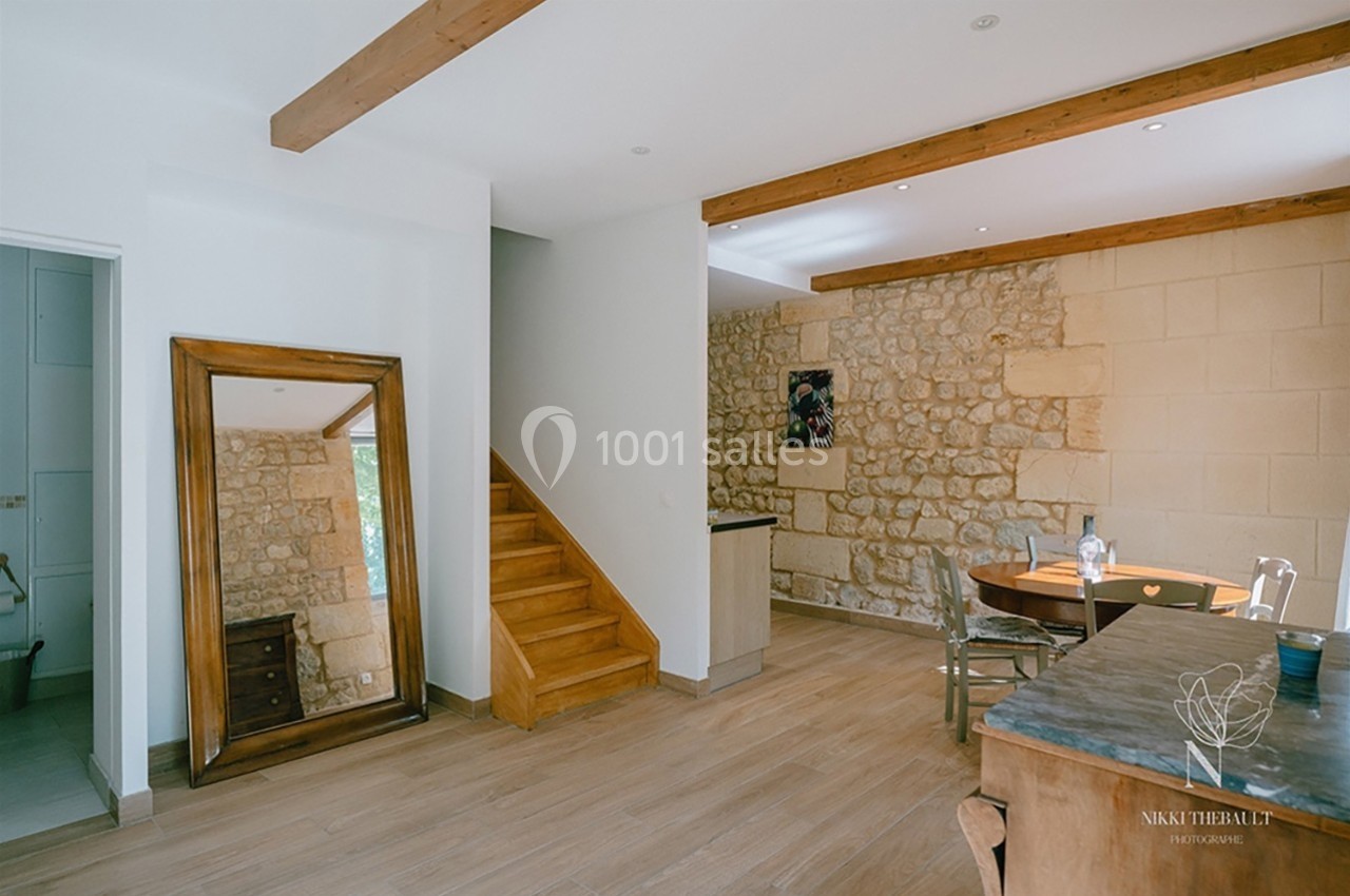 Salle à manger lumineuse avec murs en pierre, poutres apparentes, miroir au sol et escalier en bois.