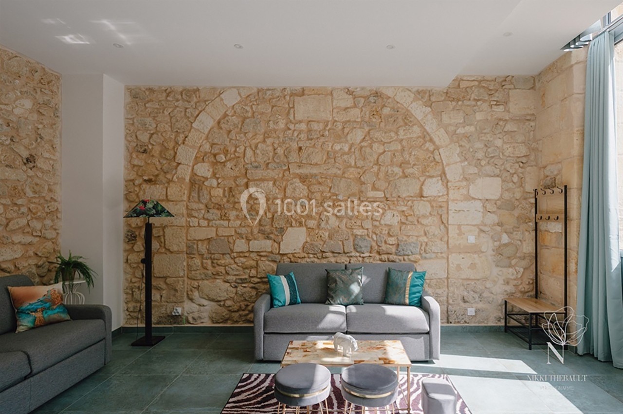 Salon lumineux avec murs en pierre apparente, canapé gris, coussins colorés et table basse sur tapis à motifs.