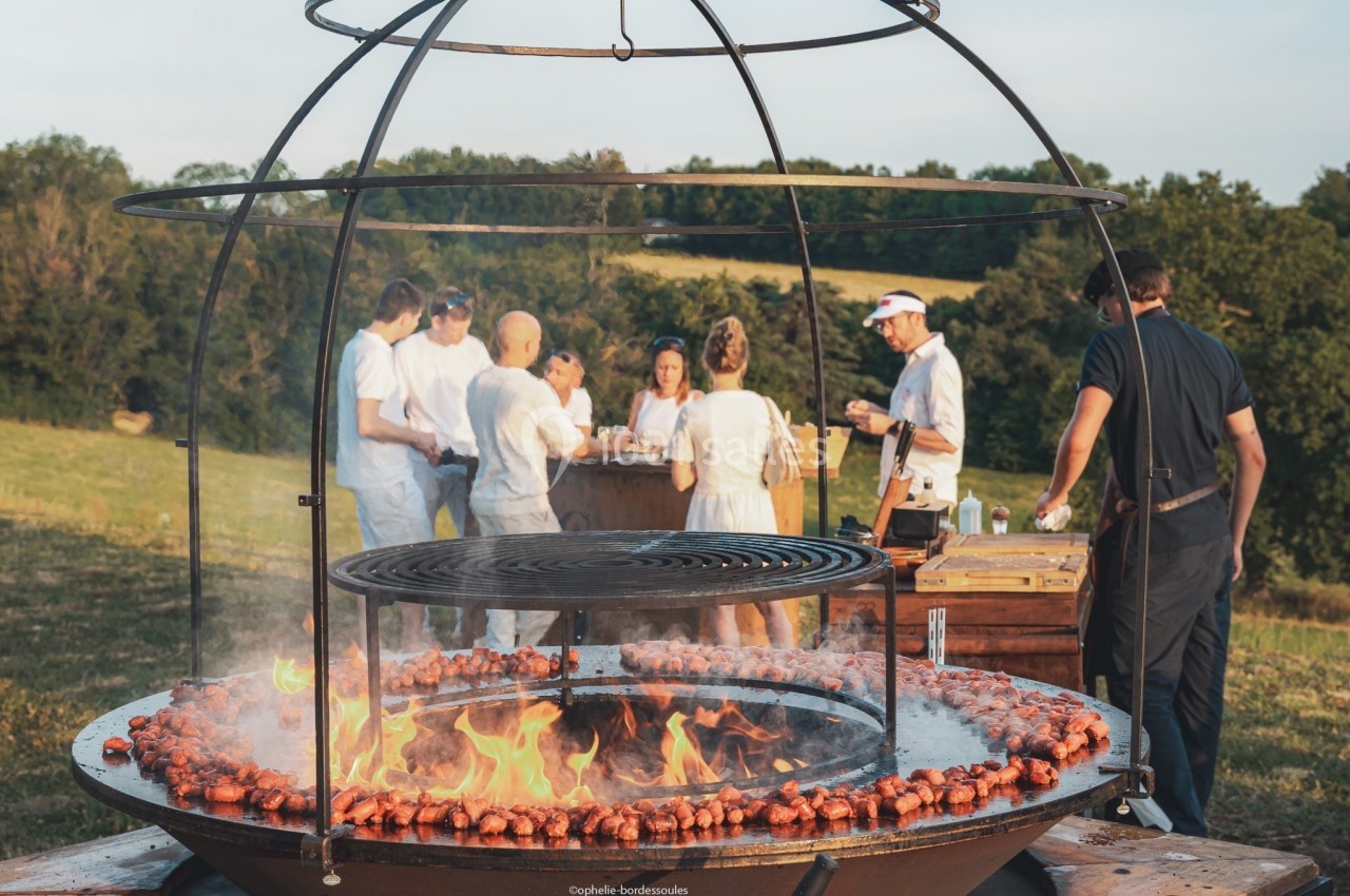Des personnes discutent autour d'un barbecue circulaire en plein air, avec des saucisses grillant sur le feu.