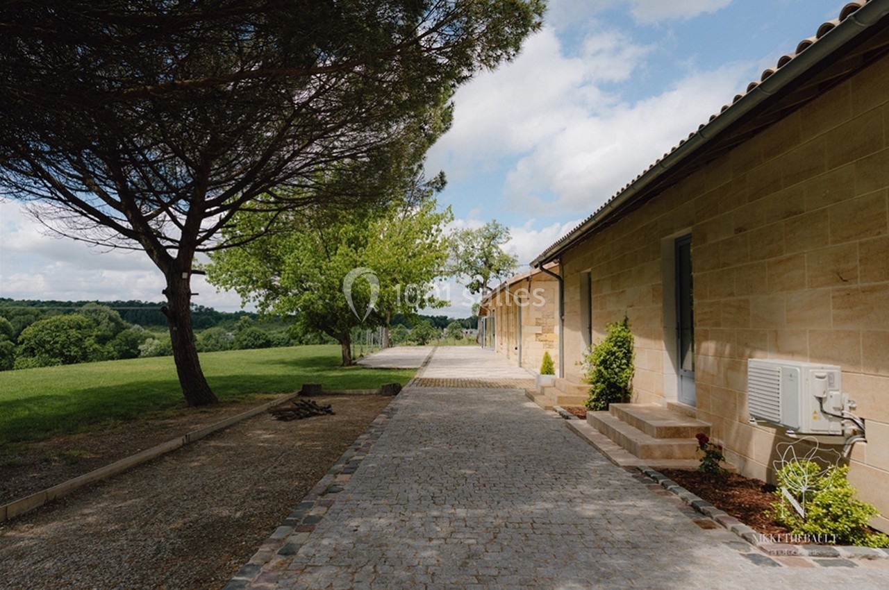 Allée pavée bordée d'arbres et d'un bâtiment en pierre, donnant sur un paysage verdoyant sous un ciel partiellement nuageux.