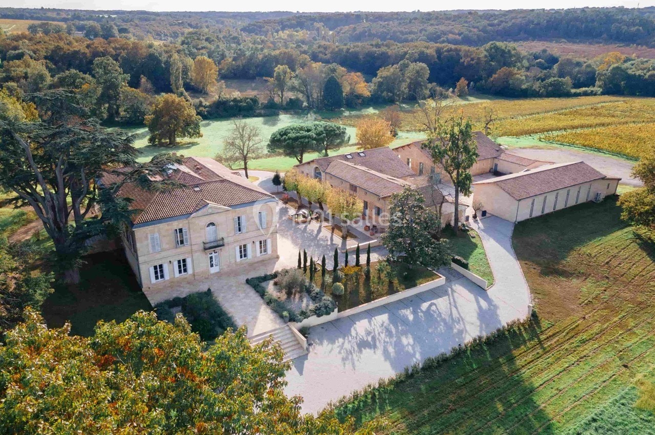 Vue aérienne d'un domaine viticole avec bâtiments en pierre, cour centrale, vignes et paysage boisé en arrière-plan.