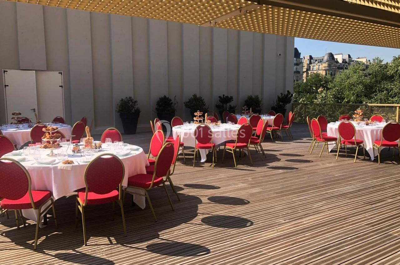 Tables rondes dressées avec nappes blanches et chaises rouges sur une terrasse en bois, entourée de verdure et bâtiments.