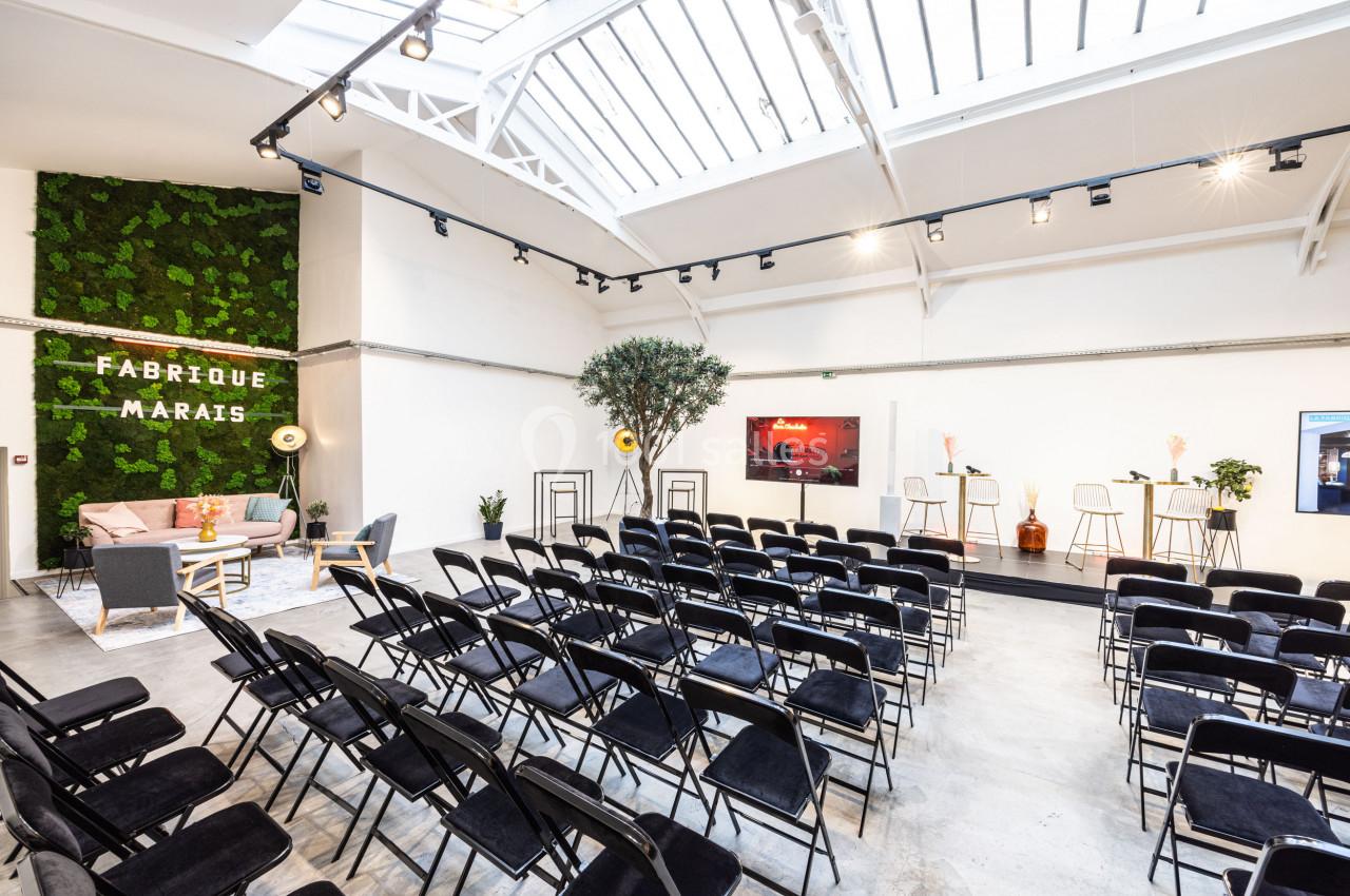 Salle lumineuse avec rangées de chaises noires, espace scénique, mur végétal et décoration moderne.