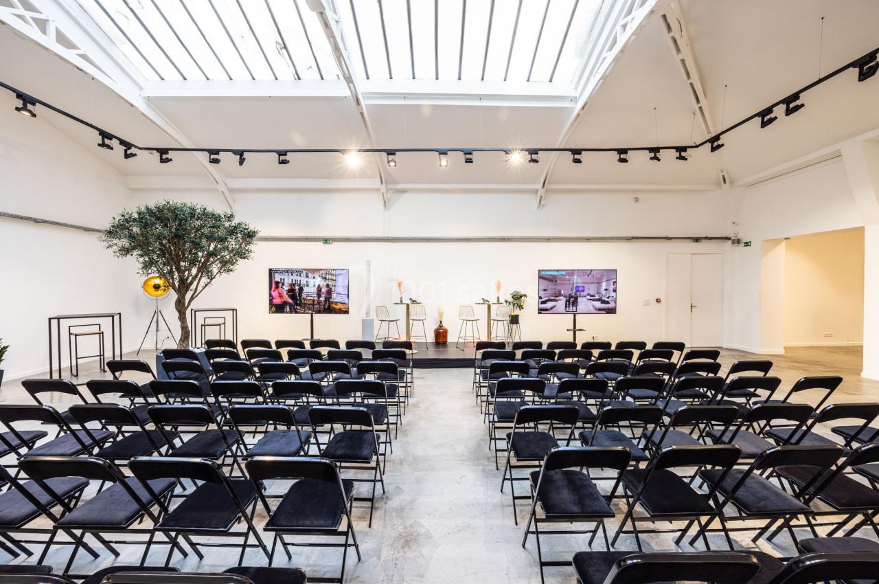 Salle lumineuse avec des rangées de chaises noires, un podium central et deux écrans sous une verrière.