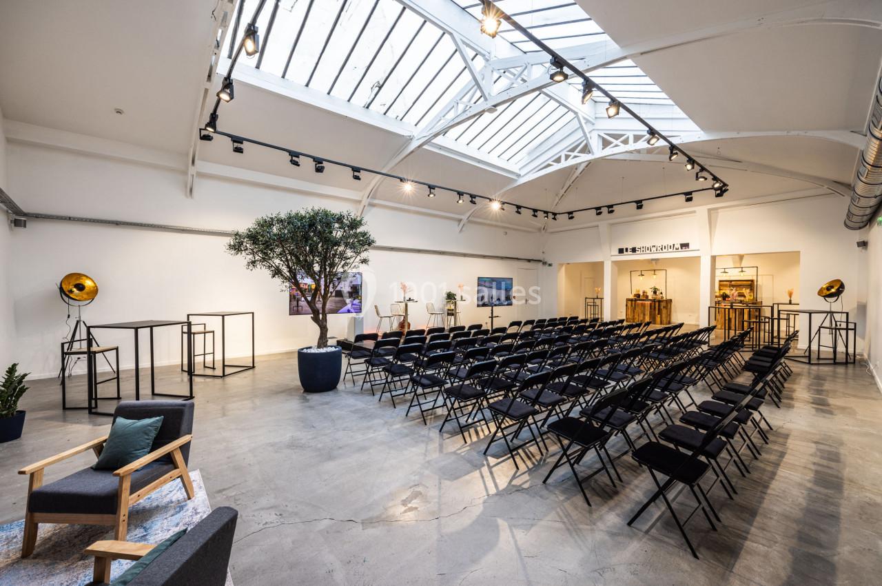 Salle lumineuse avec verrière, rangées de chaises noires, un olivier en pot et un espace aménagé en fond.