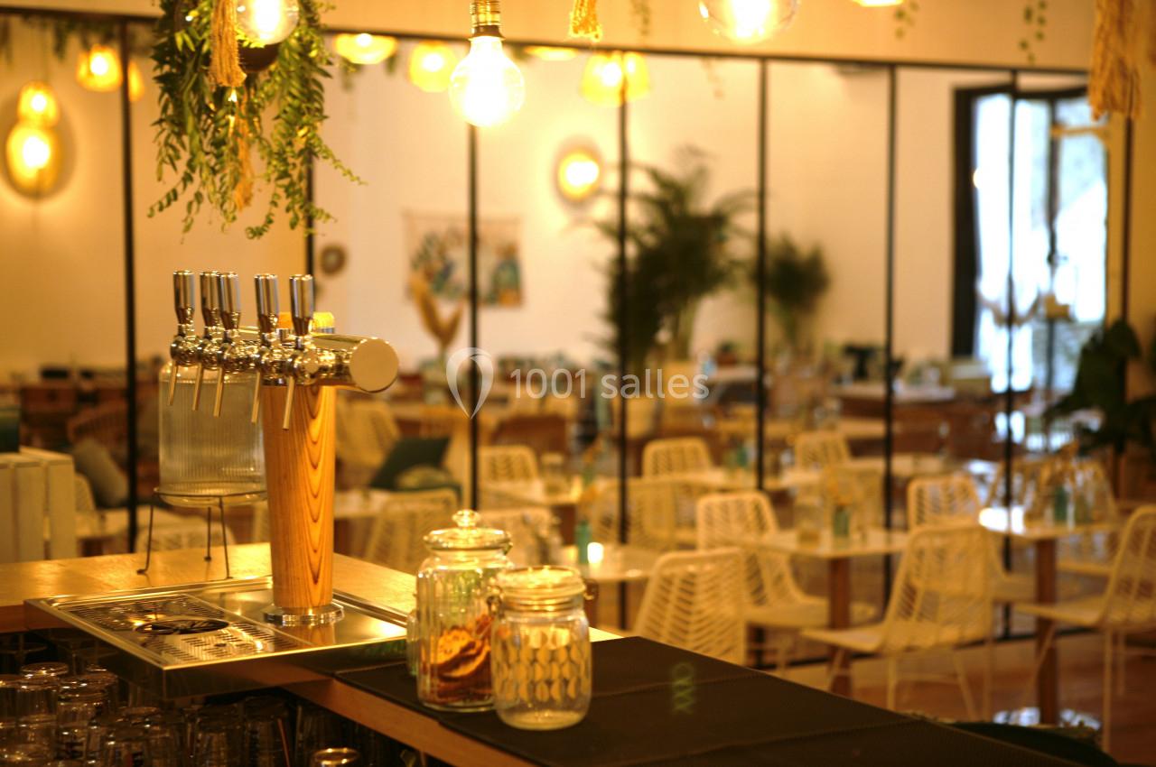 Comptoir avec tireuses à bière et bocaux, devant une salle lumineuse aménagée avec des tables et des chaises en rotin.