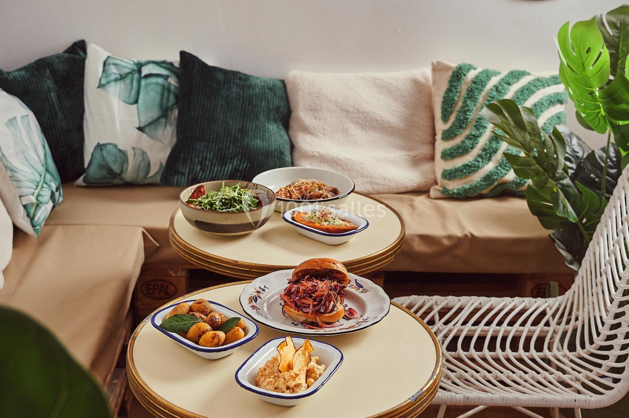 Assiettes de plats variés disposées sur des tables rondes dans un espace cosy avec coussins et décorations murales…