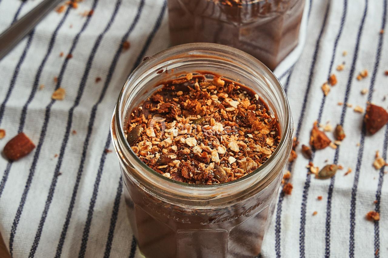 Deux pots de dessert au chocolat garnis de granola, posés sur un torchon rayé avec des cuillères à côté.